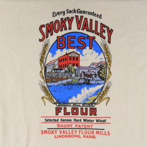 On cream colored fabric, there's a reproduction of the graphic design of a flour bag. In the center oval matte, there's a three-story brick mill across a river, surrounded by industrial buildings. Around the oval are several stalks of wheat. Below the image it's labeled "Smoky Hill River." Wording reads: "Every Sack Guaranteed. Smoky Valley Best Flour. Selected Kansas Hard Winter Wheat. Short Patent. Smoky Valley Flour Mills. Lindsborg, Kans."