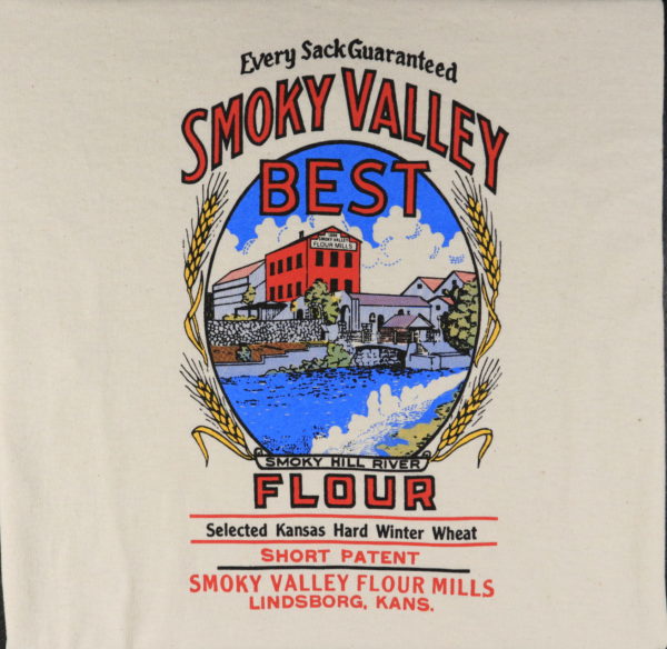 On cream colored fabric, there's a reproduction of the graphic design of a flour bag. In the center oval matte, there's a three-story brick mill across a river, surrounded by industrial buildings. Around the oval are several stalks of wheat. Below the image it's labeled "Smoky Hill River." Wording reads: "Every Sack Guaranteed. Smoky Valley Best Flour. Selected Kansas Hard Winter Wheat. Short Patent. Smoky Valley Flour Mills. Lindsborg, Kans."