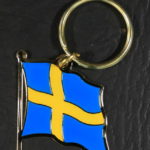 A small metal keychain in the shape of the Swedish flag, waving in the wind - a yellow cross on a light blue background.