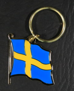 A small metal keychain in the shape of the Swedish flag, waving in the wind - a yellow cross on a light blue background.