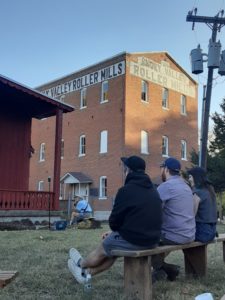 Four people on benches in front of a three story brick-walled mill.