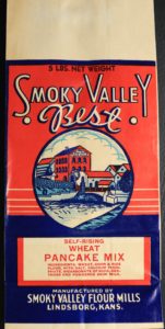 A flat, unused paper flour bag in red, white and blue. At the center is a three-story mill and industrial buildings across a dammed river. Text reads: "5 pounds net weight. Smoky Valley Best. Self-Rising Wheat Pancake Mix. Ingredients: Wheat, Corn & Rice flour, with salt, calcium phosphate, bicarbonate of soda, dextrose and powdered skim milk. Manufactured by Smoky Valley Flour Mills. Lindsborg, Kansas."