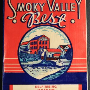 A flat, unused paper flour bag in red, white and blue. At the center is a three-story mill and industrial buildings across a dammed river. Text reads: "5 pounds net weight. Smoky Valley Best. Self-Rising Wheat Pancake Mix. Ingredients: Wheat, Corn & Rice flour, with salt, calcium phosphate, bicarbonate of soda, dextrose and powdered skim milk. Manufactured by Smoky Valley Flour Mills. Lindsborg, Kansas."