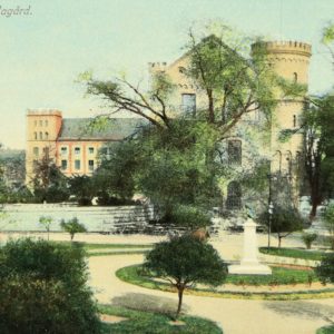 Postcard image of pale red multi story building next to a yellow brick multi story building. A circle walk way is in the foreground with a statue surrounded by green grass in its center. Trees with green leaves surround either side of the circle pathway and are in front of the yellow brick building. Text on the upper left hand corner reads Parti från Lundagård.