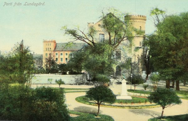 Postcard image of pale red multi story building next to a yellow brick multi story building. A circle walk way is in the foreground with a statue surrounded by green grass in its center. Trees with green leaves surround either side of the circle pathway and are in front of the yellow brick building. Text on the upper left hand corner reads Parti från Lundagård.