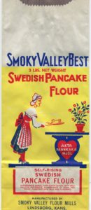 A flat, unused paper flour bag in yellow, red, white, blue and green. A woman in traditional Swedish dress places a stack of pancakes onto a table supported by a heart-shaped base and flowers in a pot resting on top of it. Text reads: "Smoky Valley Best. 3 pounds net weight. Swedish Pancake Flour. Akta Pannkaka Mjol. Self-Rising Swedish Pancake Flour. Ingredients: Wheat, corn & rice flour with salt, calcium phosphate, bicarbonate of soda, dextrose, powdered skim milk, and flavoring. Manufactured by Smoky Valley Flour Mills. Lindsborg, Kansas."