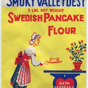 A flat, unused paper flour bag in yellow, red, white, blue and green. A woman in traditional Swedish dress places a stack of pancakes onto a table supported by a heart-shaped base and flowers in a pot resting on top of it. Text reads: "Smoky Valley Best. 3 pounds net weight. Swedish Pancake Flour. Akta Pannkaka Mjol. Self-Rising Swedish Pancake Flour. Ingredients: Wheat, corn & rice flour with salt, calcium phosphate, bicarbonate of soda, dextrose, powdered skim milk, and flavoring. Manufactured by Smoky Valley Flour Mills. Lindsborg, Kansas."