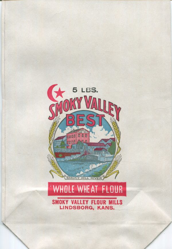 A large, flat unused flour bag in full color. At the center in an oval frame is a view of a red brick, three-story mill building with a gabled roof, surrounded by lower level industrial buildings. In the foreground is a blue river that has been dammed. On either side of the frame are stalks of yellow wheat. The label at the bottom reads "Smoky Hill River." To the upper left is a red crescent moon and star. Text on the bag reads: "5 pounds. Smoky Valley Best. Whole Wheat Flour. Smoky Valley Flour Mills. Lindsborg, Kansas."