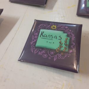 On a square button, a light teal shape labeled "Kansas" and with cute smile is on a purple background. Around the state are wheat stalks, a sunflower, and a number of doodle sketches.