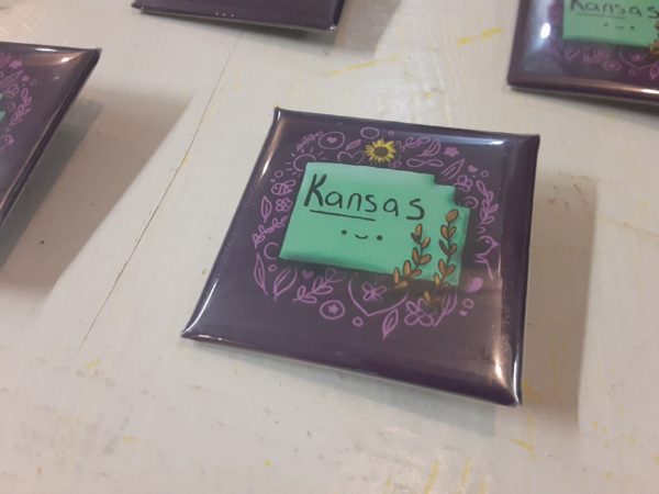 On a square button, a light teal shape labeled "Kansas" and with cute smile is on a purple background. Around the state are wheat stalks, a sunflower, and a number of doodle sketches.