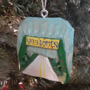 A hand-carved wooden Christmas ornament hanging in front of an evergreen tree in close-up. The roughly square ornament features a green frame bridge and a gray road over it against a light blue sky. The dark green bridge has small red Christmas lights lining it and a yellow sign reading "Valkommen" is above the entrance on the bridge.