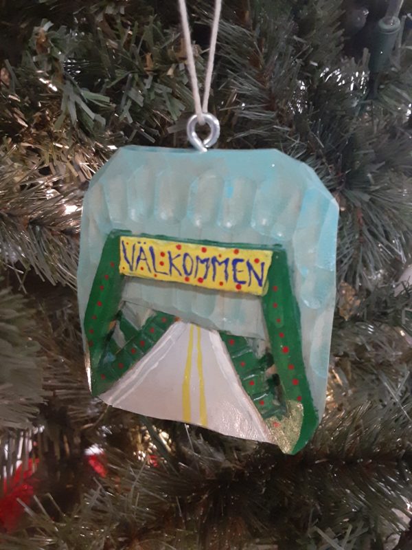 A hand-carved wooden Christmas ornament hanging in front of an evergreen tree in close-up. The roughly square ornament features a green frame bridge and a gray road over it against a light blue sky. The dark green bridge has small red Christmas lights lining it and a yellow sign reading "Valkommen" is above the entrance on the bridge.