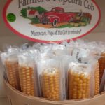 A basket of yellow corn cobs individually wrapped in clear plastic. A sign at the back of the basket pictures an old fashioned red pickup on a farm and a farmer in overalls stacking baskets of produce. Wording reads: All-Natural Farmer's Popcorn Cob Microwave. Pops off the Cob in minutes!