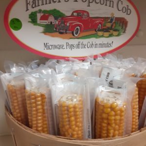 A basket of yellow corn cobs individually wrapped in clear plastic. A sign at the back of the basket pictures an old fashioned red pickup on a farm and a farmer in overalls stacking baskets of produce. Wording reads: All-Natural Farmer's Popcorn Cob Microwave. Pops off the Cob in minutes!