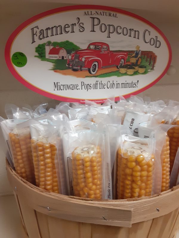 A basket of yellow corn cobs individually wrapped in clear plastic. A sign at the back of the basket pictures an old fashioned red pickup on a farm and a farmer in overalls stacking baskets of produce. Wording reads: All-Natural Farmer's Popcorn Cob Microwave. Pops off the Cob in minutes!