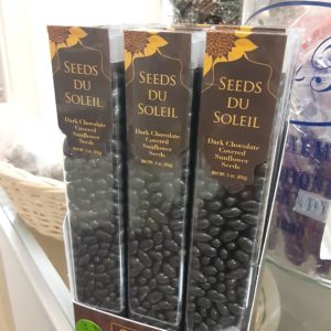A box filled with tall, clear plastic containers of dark brown small candy. They have dark brown labels as well featuring sunflowers. Written on the boxes: Seeds Du Soleil Dark Chocolate Covered Sunflower Seeds Net Wt. 3 oz. (85g)