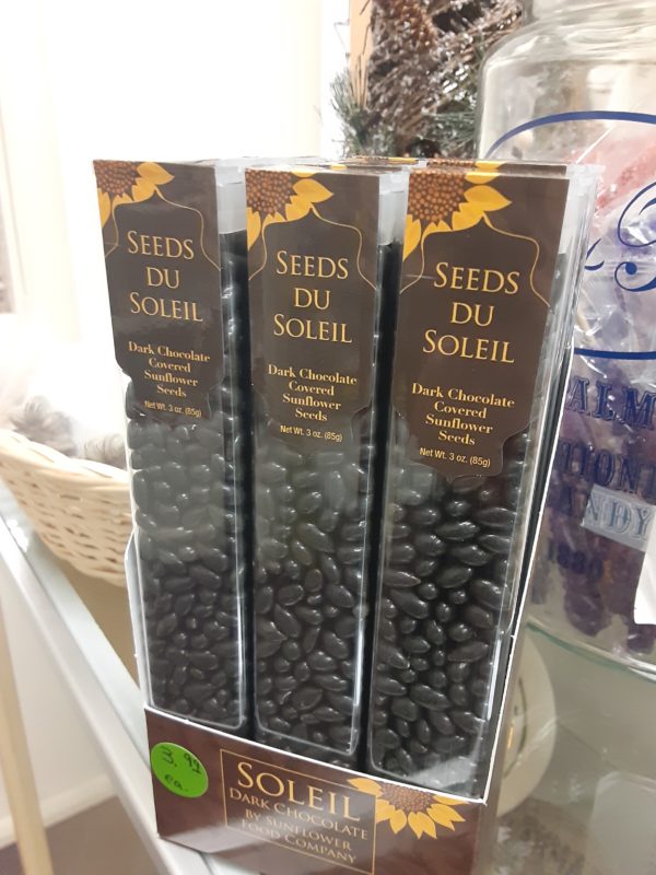 A box filled with tall, clear plastic containers of dark brown small candy. They have dark brown labels as well featuring sunflowers. Written on the boxes: Seeds Du Soleil Dark Chocolate Covered Sunflower Seeds Net Wt. 3 oz. (85g)