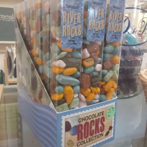A light blue box filled with tall, clear plastic containers of multicolored small candy with the appearance of smooth river rocks. Written on the box: Chocolate Rocks Collection Written on the individual boxes: Chocolate River Rocks Net Wt. 2.8 oz. (85g)