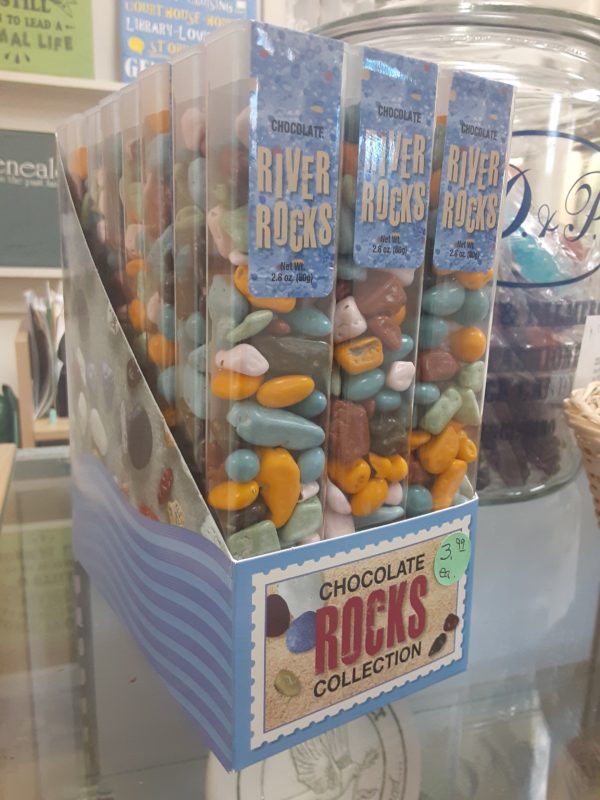 A light blue box filled with tall, clear plastic containers of multicolored small candy with the appearance of smooth river rocks. Written on the box: Chocolate Rocks Collection Written on the individual boxes: Chocolate River Rocks Net Wt. 2.8 oz. (85g)