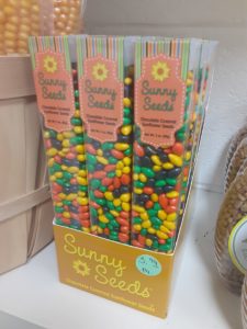 A box filled with tall, clear plastic containers of multicolored small candy. Written on the boxes: Sunny Seeds Chocolate Covered Sunflower Seeds Net Wt. 3 oz. (85g)