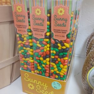 A box filled with tall, clear plastic containers of multicolored small candy. Written on the boxes: Sunny Seeds Chocolate Covered Sunflower Seeds Net Wt. 3 oz. (85g)