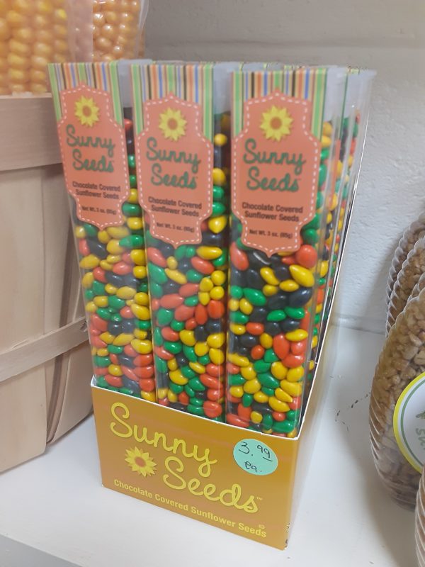 A box filled with tall, clear plastic containers of multicolored small candy. Written on the boxes: Sunny Seeds Chocolate Covered Sunflower Seeds Net Wt. 3 oz. (85g)