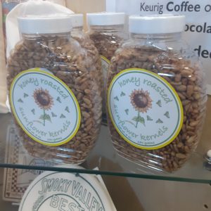 Several clear plastic containers shaped like a round honey container and filled with shelled sunflower seeds. A round sticker on the front has a sunflower surrounded by butterflies. Writing reads: Honey roasted sunflower kernels Net wt. 6 oz. 170 grams