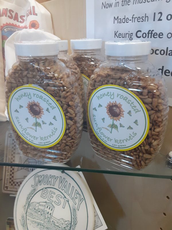 Several clear plastic containers shaped like a round honey container and filled with shelled sunflower seeds. A round sticker on the front has a sunflower surrounded by butterflies. Writing reads: Honey roasted sunflower kernels Net wt. 6 oz. 170 grams