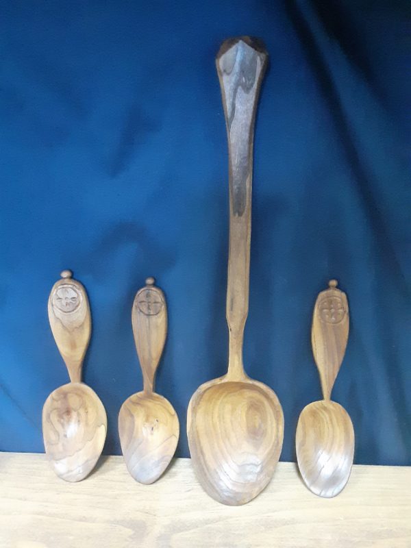 Four wooden spoons on a navy blue background. One is much longer than the other three, with a tapered handle and a large bowl. The other three smaller spoons have a small symmetrical cross design surrounded by a circle. The bowl of these spoons is more oval and shallow, with handles that are broad at the end, but thin to a narrow neck at the bowl.