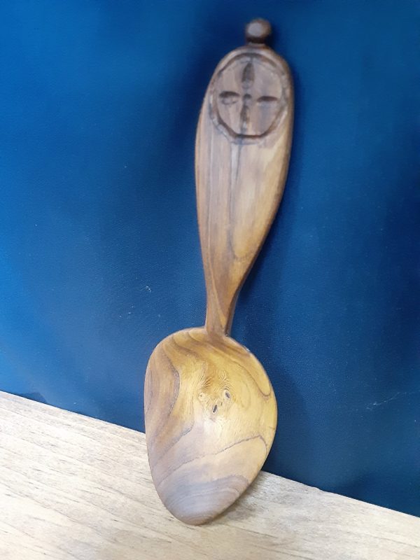A small natural-color wooden spoon on a navy blue fabric background. It has a small symmetrical cross design surrounded by a circle. The bowl of the spoon is an elongated oval and shallow, with a handle broad at the end, but thinning to a narrow neck at the bowl.