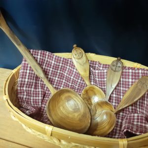 Four wooden spoons layered in a light colored natural woven basket on red gingham fabric. The basket is on a wooden shelf on a navy blue fabric background. One spoon is much longer than the other three, with a tapered handle and a large bowl. The other three smaller spoons have a small symmetrical cross design surrounded by a circle. The bowl of these spoons is more oval and shallow, with handles that are broad at the end, but thin to a narrow neck at the bowl.