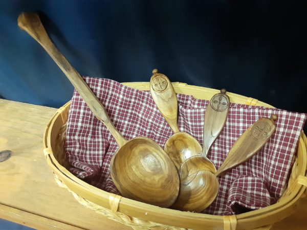 Four wooden spoons layered in a light colored natural woven basket on red gingham fabric. The basket is on a wooden shelf on a navy blue fabric background. One spoon is much longer than the other three, with a tapered handle and a large bowl. The other three smaller spoons have a small symmetrical cross design surrounded by a circle. The bowl of these spoons is more oval and shallow, with handles that are broad at the end, but thin to a narrow neck at the bowl.