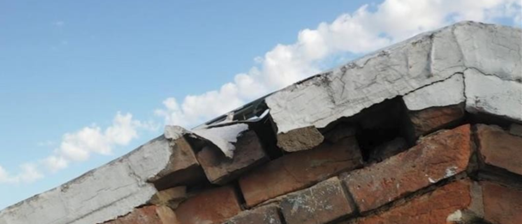 The peak of a brick building at the roof line with blue sky and clouds in the background. Several bricks are missing just under the roof line.